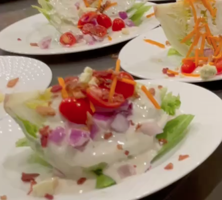 Wedge salad topped with sliced cherry tomatoes, shredded carrot, diced red onion, bacon bits and ranch dressing.